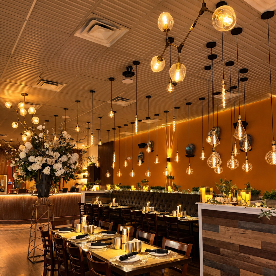 Elegant restaurant dining room with warmly lit pendant bulbs, set tables, tufted seating, orange walls, and a large floral arrangement.