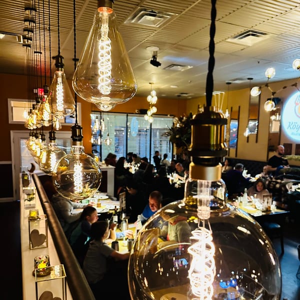 Warmly lit Koy Grill dining room with Edison bulbs in the foreground and blurred patrons.