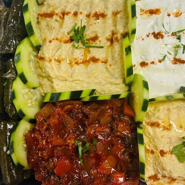 A Mediterranean mezze platter with hummus, tzatziki, dolmades, and a tomato relish, garnished with cucumber slices.