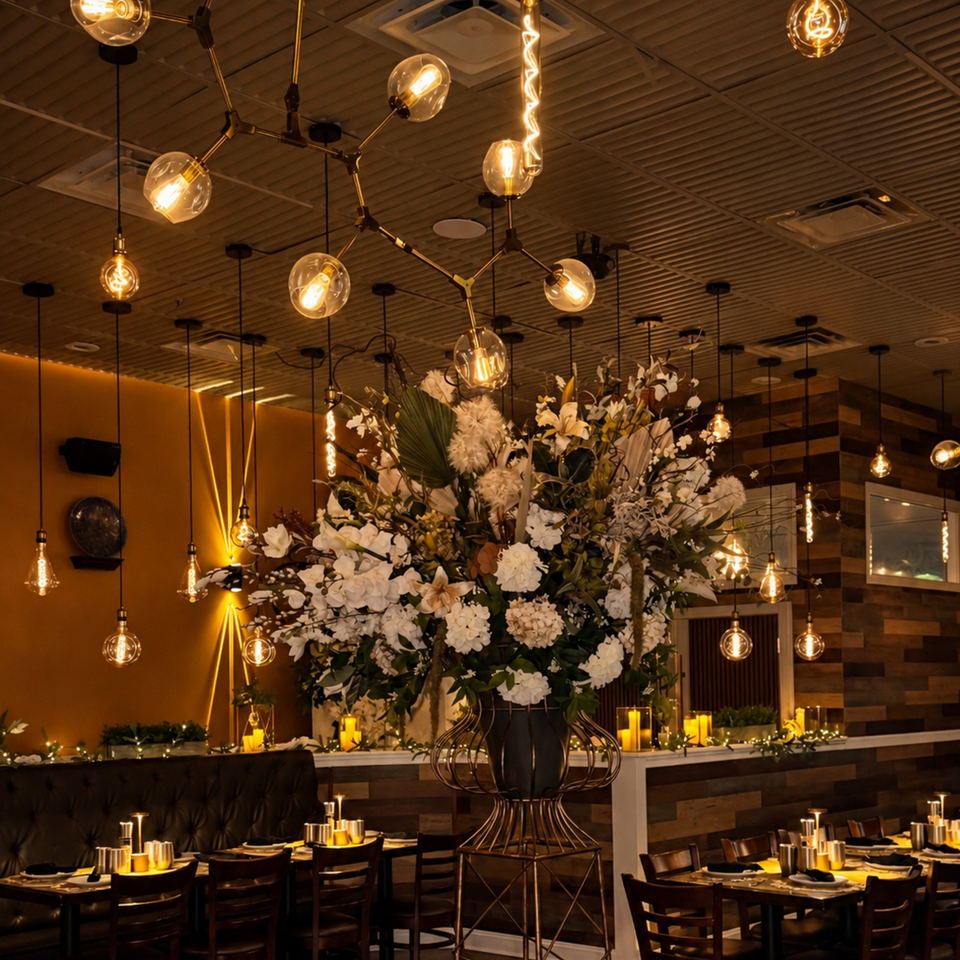 Upscale restaurant dining room with glowing pendant lights, wood accents, set tables, and a large white floral centerpiece.