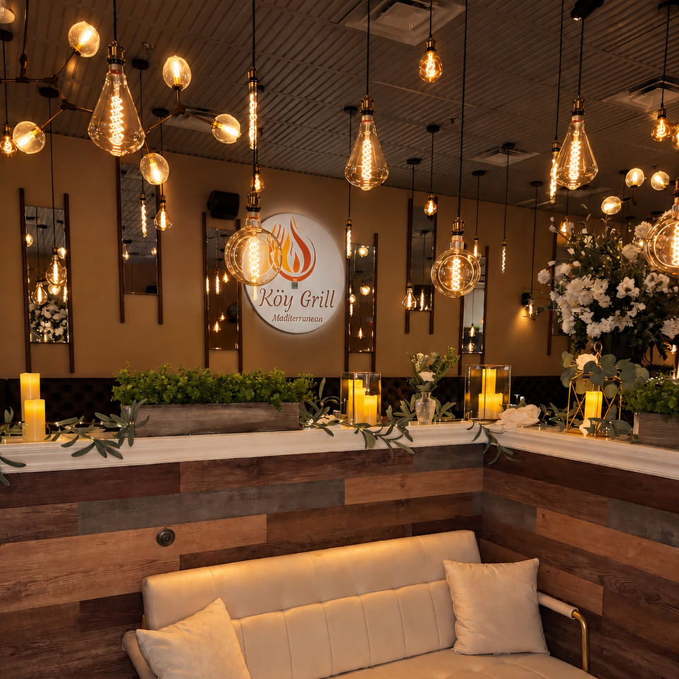 Warmly lit Kōy Grill Mediterranean restaurant interior with hanging Edison bulbs, candles, floral arrangements, and a cushioned booth.