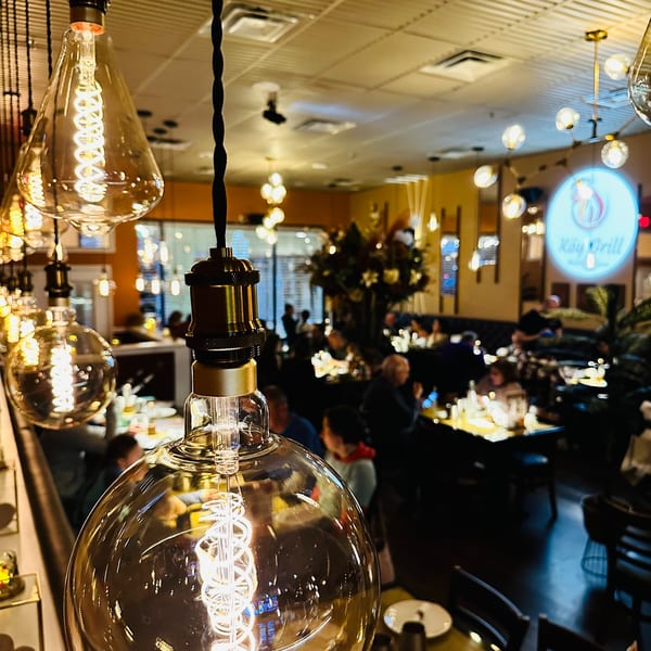 Warmly lit Koy Grill dining room with Edison bulbs in the foreground and blurred diners.