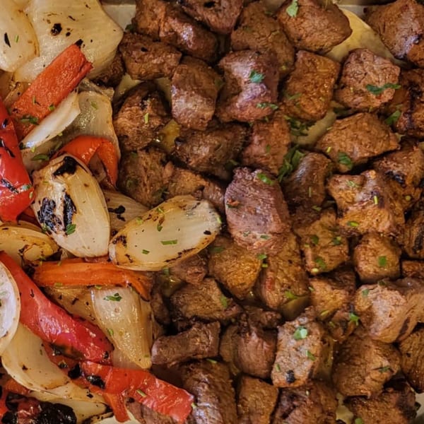 Grilled beef chunks with charred onions and red bell peppers, garnished with herbs.