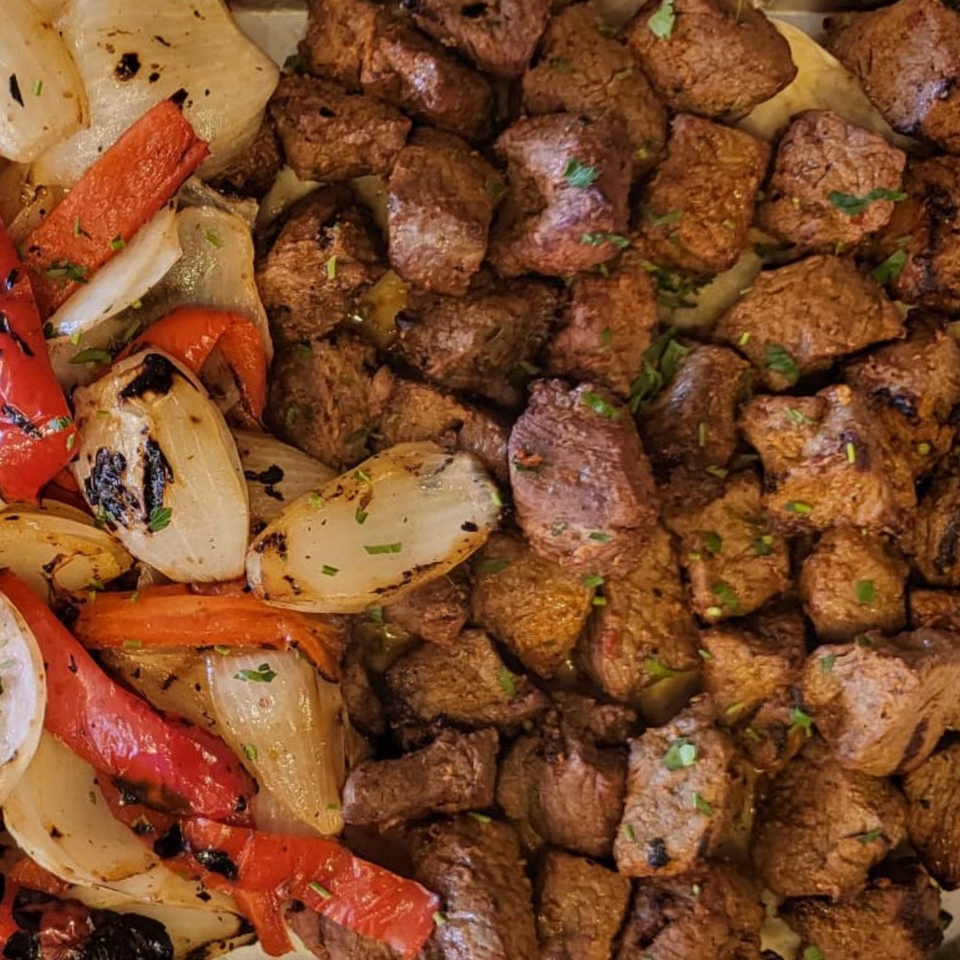 Grilled beef chunks with charred onions and red bell peppers, garnished with herbs.