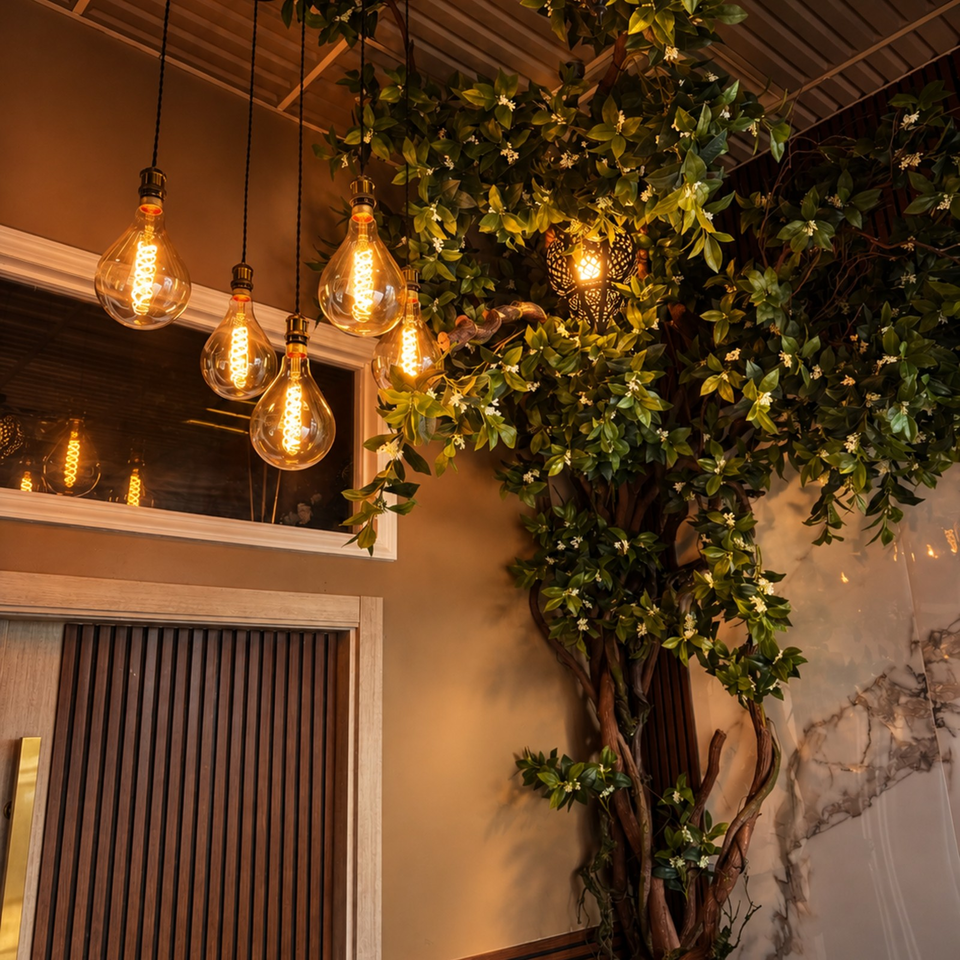 Warmly lit restaurant interior with hanging Edison bulbs, a leafy flowering tree, wood paneling, and marble accents.