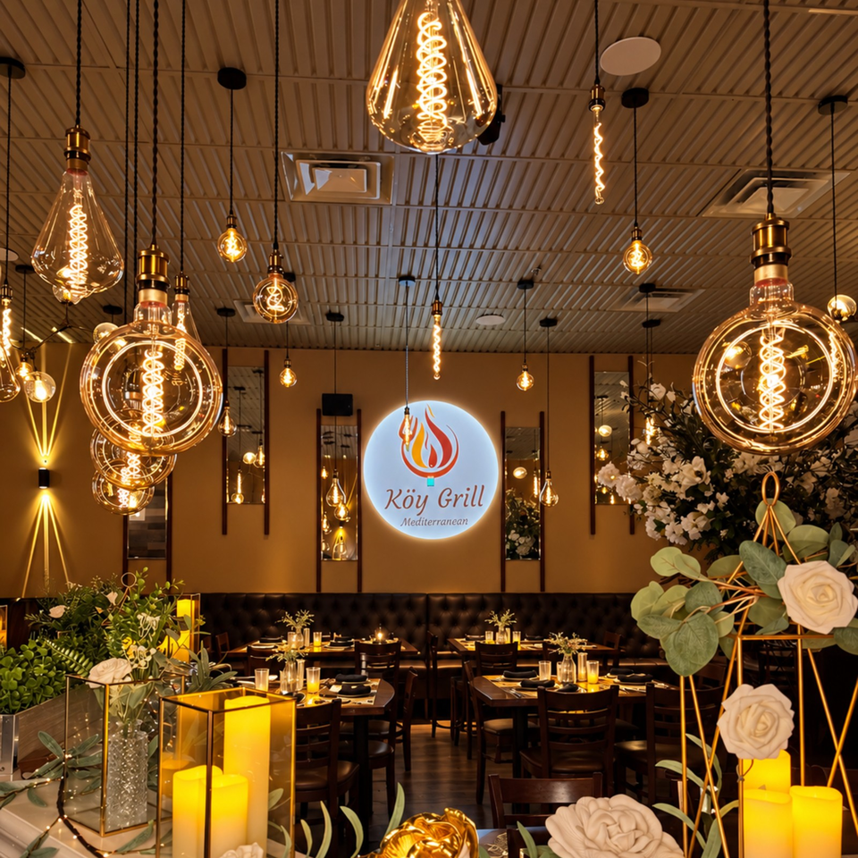Warmly lit Köy Grill Mediterranean restaurant interior with glowing pendant lights, candlelit tables, floral arrangements, and illuminated wall logo.
